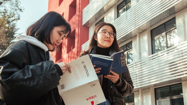 Two young women reading books outside a modern building, enjoying sunny weather.