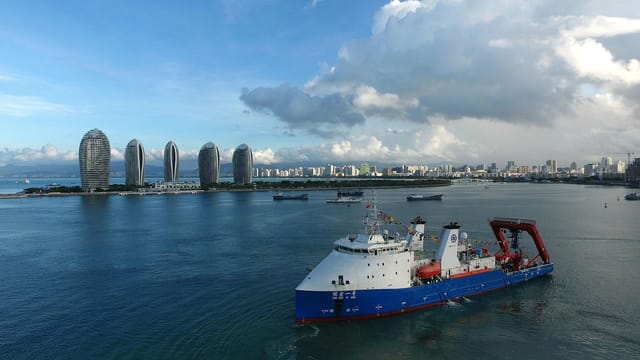 Aerial shot of a research vessel sailing near Phoenix Island, Sanya, with modern skyscrapers in the background.