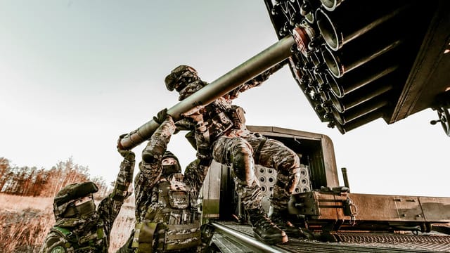 Soldiers in camouflage uniforms loading artillery equipment during a military field operation.