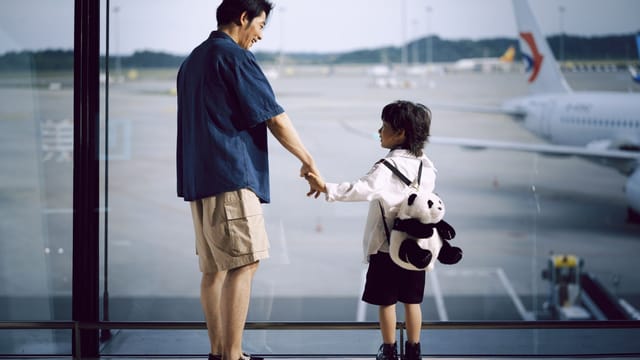 A father and child holding hands, gazing at planes through an airport window.