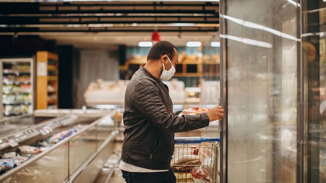 A man wearing a mask shops in a supermarket, selecting items from a refrigerated section.