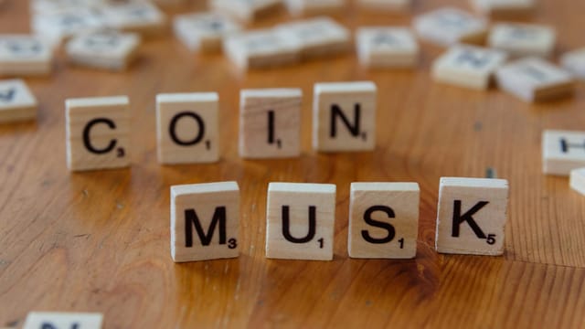 Scrabble tiles forming the words 'COIN' and 'MUSK' on a wooden table surface.