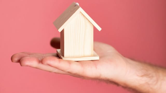 Wooden house model in hand symbolizing real estate concepts on pink background.