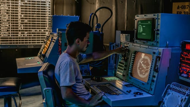 A man working at control panels in a dimly lit industrial room with various monitors.