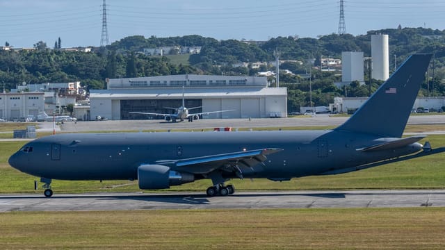 A military aircraft taxiing on a runway at an airbase surrounded by lush green hills.