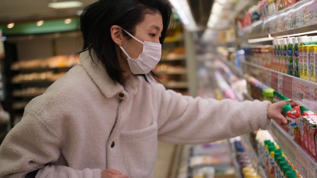 A woman wearing a face mask selects products in a supermarket beverage aisle.