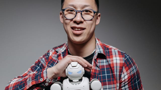 Asian man smiling, holding a small toy robot during a studio shoot. Indoors, with eyeglasses and plaid shirt.