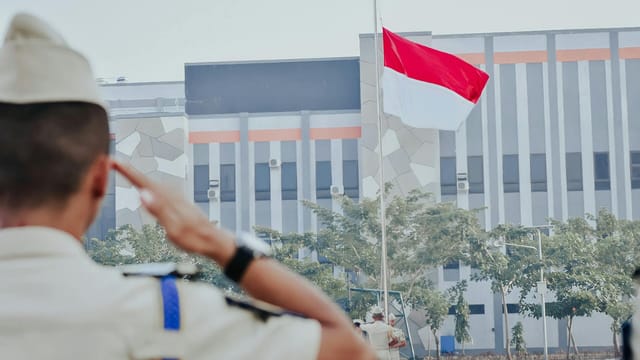 A soldier in uniform saluting the Indonesian flag during a flag raising ceremony outdoors.