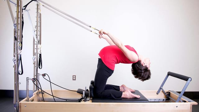 Woman performing stretching exercise on a Pilates reformer in a brightly lit gym environment.