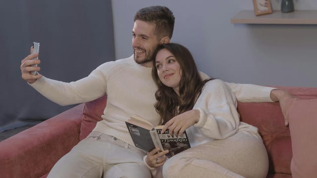 A couple relaxing on a sofa, sharing a cozy moment while taking a selfie with a smartphone.