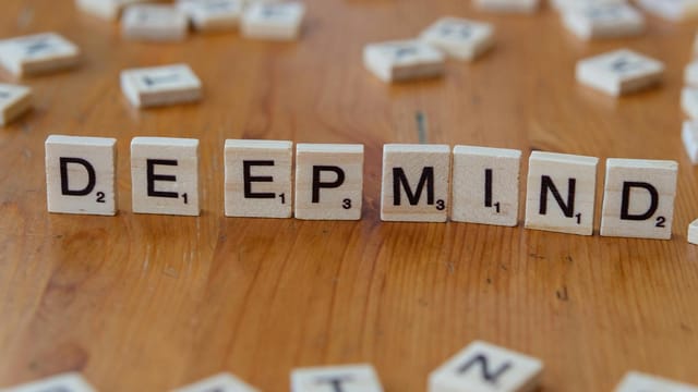 Close-up of Scrabble tiles spelling 'DeepMind' arranged on a wooden surface, with scattered tiles in the background.