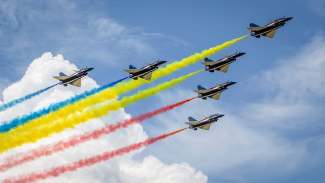 Jets performing an aerial show with colorful vapor trails against a blue sky with clouds.