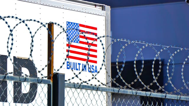 Industrial scene with barbed wire fence and American flag logo on building.