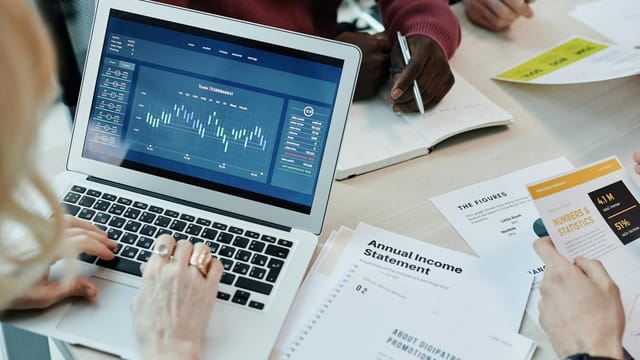 Business professionals analyzing financial data on a laptop during a meeting. Charts and reports visible.