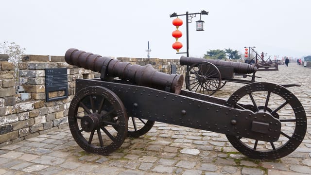 Rustic ancient cannons displayed on a historic Chinese fortress under a cloudy sky.