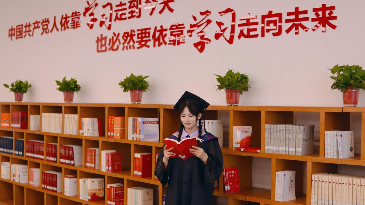 Female graduate in academic robe reading in a library with motivational Chinese text on the wall.