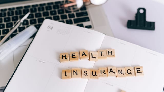 Close-up of Scrabble tiles spelling 'Health Insurance' on a planner with a laptop.