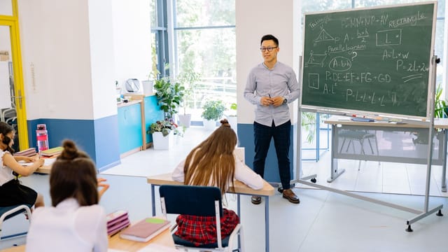 Teacher instructing students in a bright classroom with a blackboard filled with math equations.