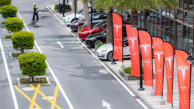 Aerial view of multiple Tesla cars parked alongside branded flags in a city street.