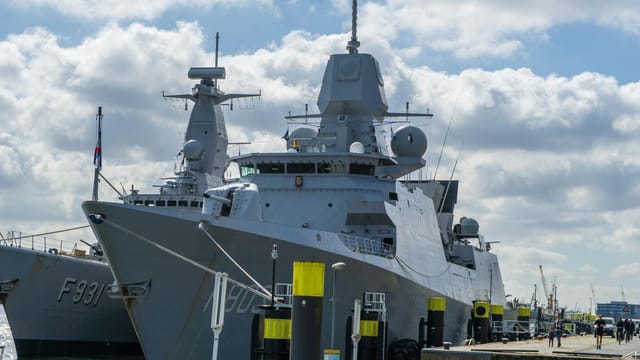 A modern naval warship moored at the port of Rotterdam under a blue sky with clouds.