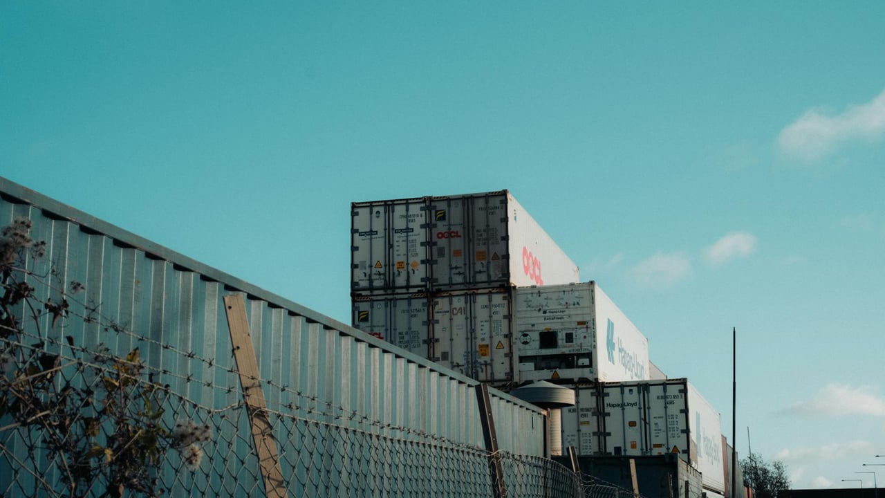 A view of stacked shipping containers beside a fence under a clear blue sky, captured outdoors.