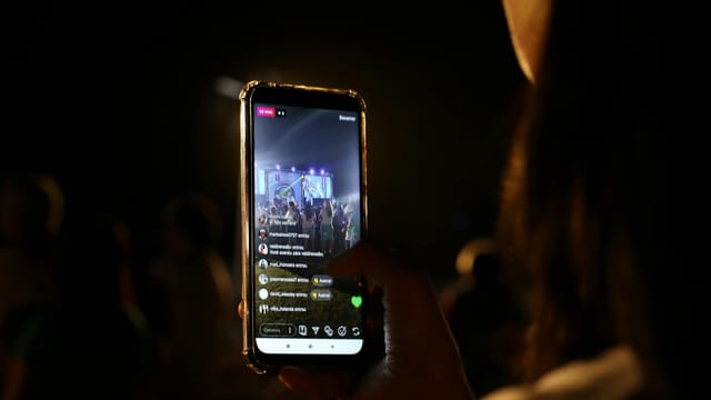 A woman live streams a concert using her smartphone at night. Vibrant stage in view.