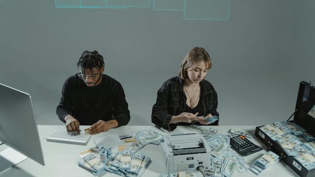 Two people counting money at a desk with computers and cash equipment.