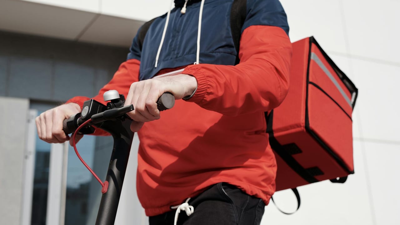A food delivery person rides a scooter outdoors, wearing a red jacket and carrying a delivery bag.