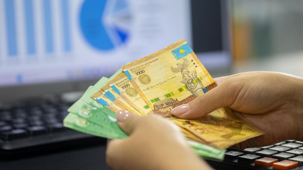 Human hands counting Kazakh tenge bills near a laptop displaying financial graphs.