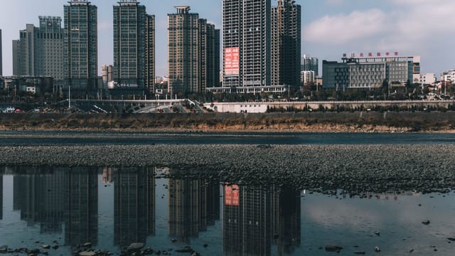 Serene cityscape with buildings reflecting in a calm river under a clear sky.