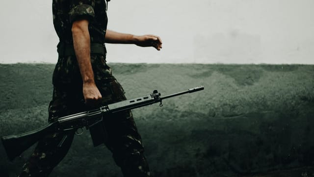 A soldier in camouflage walking with a rifle against a textured wall, day.