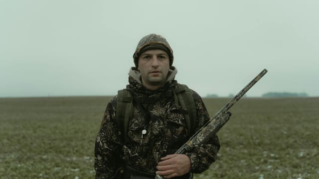 A camouflaged hunter with a rifle stands prepared outdoors during a cold season.