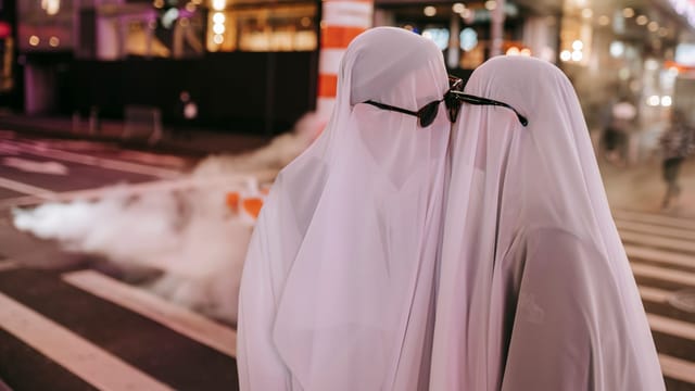 Side view of romantic anonymous couple wearing ghost costumes and sunglasses caressing each other while standing on pedestrian crossing in street with steam pipe and building on background at night time