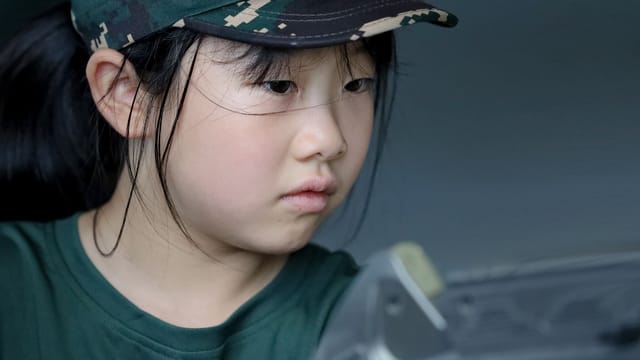 A young girl intensely focused, wearing a camouflage cap and green shirt indoors.