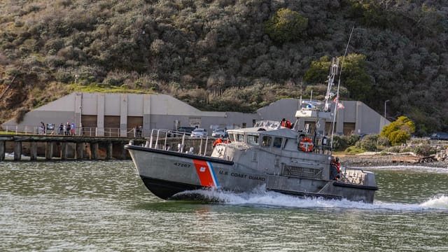 Operational U.S. Coast Guard boat cruising near seashore facility in daylight.