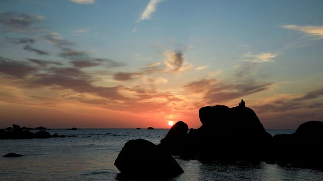 A serene sunset view over the sea with a solitary silhouette on rocks in Bretagne, France.