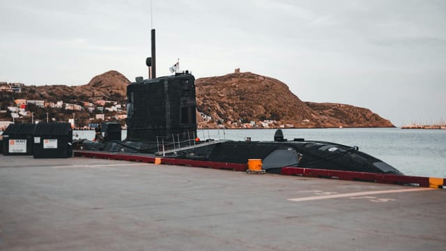 A submarine docked at an urban waterfront with scenic hills in the background.