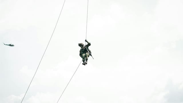 A soldier rappels from a helicopter against a cloudy sky, showcasing military training.