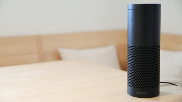 A black smart speaker resting on a light-colored wooden table in a cozy indoor setting.