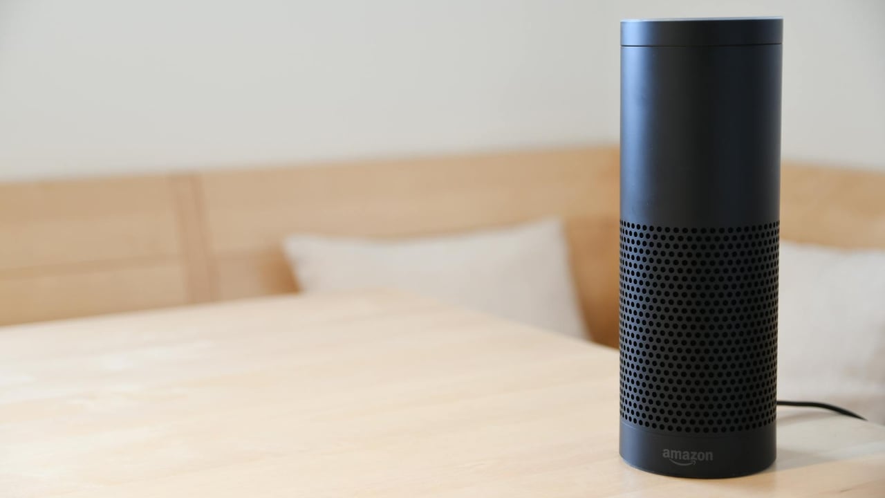 A black smart speaker resting on a light-colored wooden table in a cozy indoor setting.