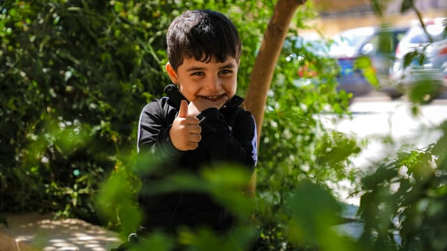 Young boy smiling and giving thumbs up outdoors amidst lush green foliage.