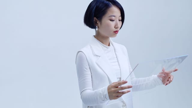 Asian woman in white blazer holding transparent glass interface on a plain background.