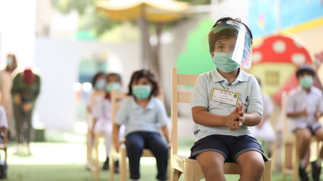 Children sitting in a classroom wearing face masks and shields for safety.