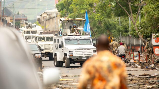 UN armored vehicles and peacekeepers patrol a busy urban street.