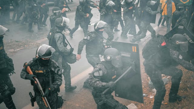 Riot police with shields confront protesters in a smoky urban setting.