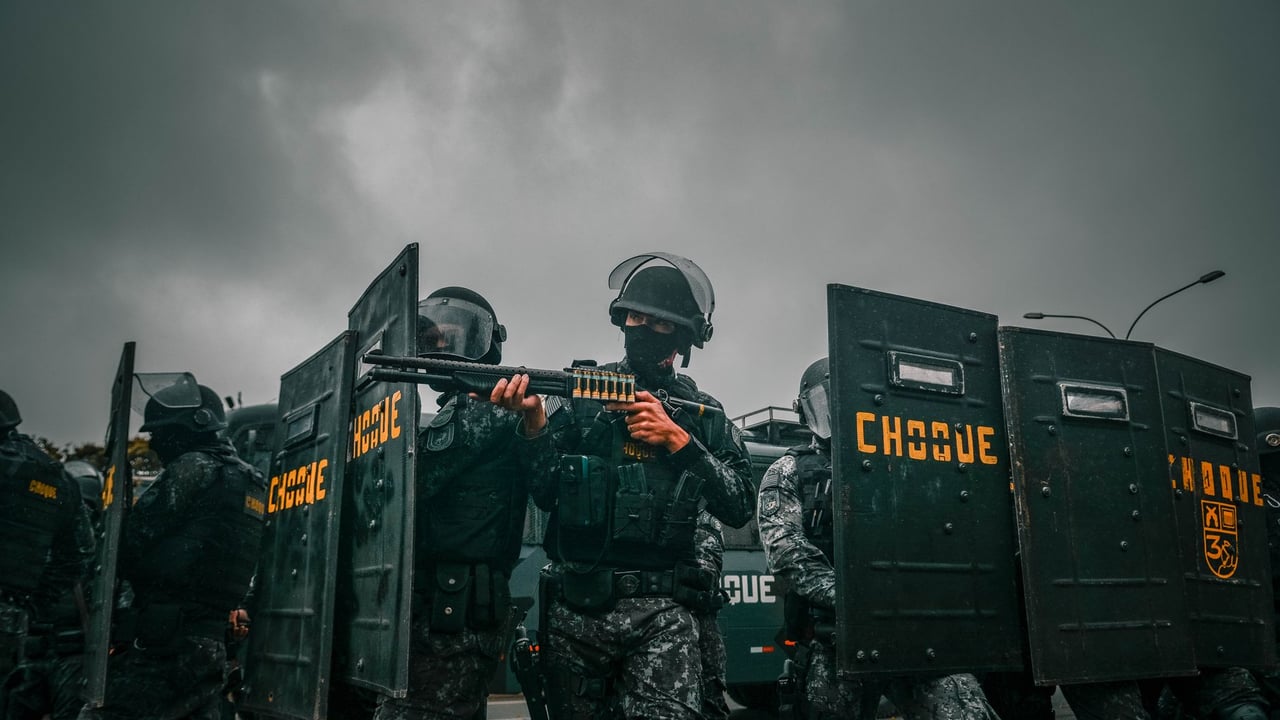 Riot police in tactical gear holding shields during a stormy protest.