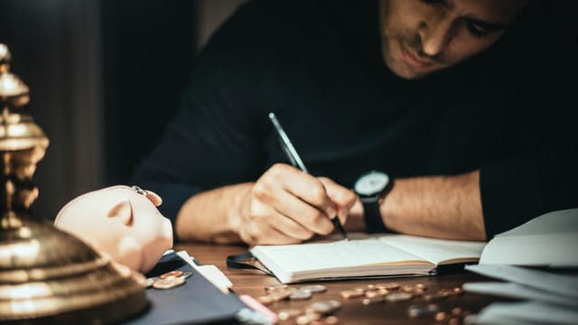 Crop elegant man taking notes in journal while working at desk with coins and piggybank in lamplight