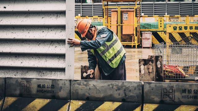 A construction worker in a reflective vest and hard hat at a busy urban construction site.