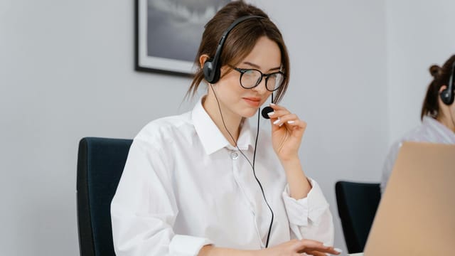 Young woman in glasses and headset providing customer support at a laptop in an office setting.