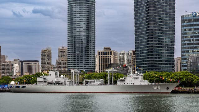 Military ship docked alongside a modern city skyline with high-rise buildings.